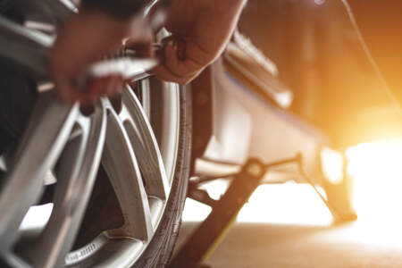 Nahaufnahme: Eine Person löst Radmuttern mit einem Werkzeug, während das Auto mit einem Wagenheber angehoben ist; warmes Sonnenlicht im Hintergrund.