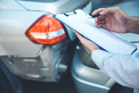 Nahaufnahme: Eine Person hält ein Klemmbrett und einen Stift, während im Hintergrund ein beschädigtes Heck eines Autos zu sehen ist.