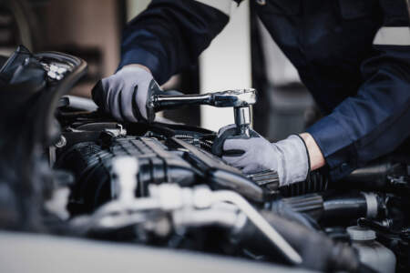 Nahaufnahme: Ein Mechaniker mit Arbeitshandschuhen nutzt eine Ratsche, um im Motorraum eines Autos eine Schraube zu lösen oder festzuziehen.