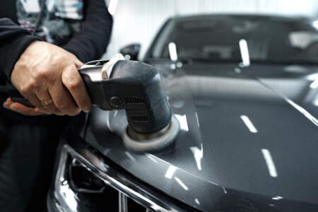 Nahaufnahme: Eine Person führt eine elektrische Poliermaschine über die Motorhaube eines dunklen Autos; Werkstatt-/Aufbereitungsumgebung im Hintergrund.