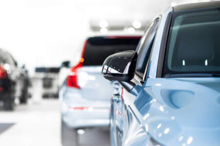Seitenansicht eines blauen Fahrzeugs mit Fokus auf den Außenspiegel; im Hintergrund stehen weitere Autos unscharf in einem hellen Showroom.