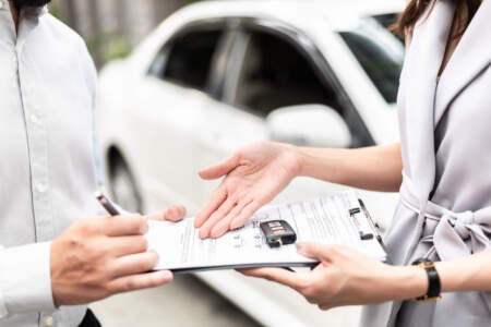 Zwei Personen besprechen ein Formular auf einem Klemmbrett; ein Autoschlüssel liegt darauf, während eine Person mit Stift notiert und die andere mit der Hand auf das Dokument zeigt. Im Hintergrund steht ein weißes Auto.